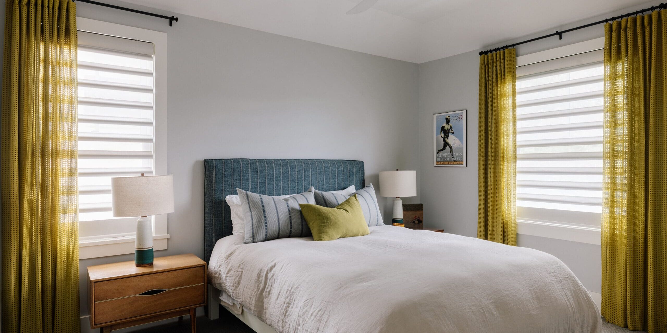 Windows Dressed Up | August Photoshoots A modern bedroom with a blue upholstered bed, white bedding, two wooden nightstands with lamps, yellow curtains, and two large windows.