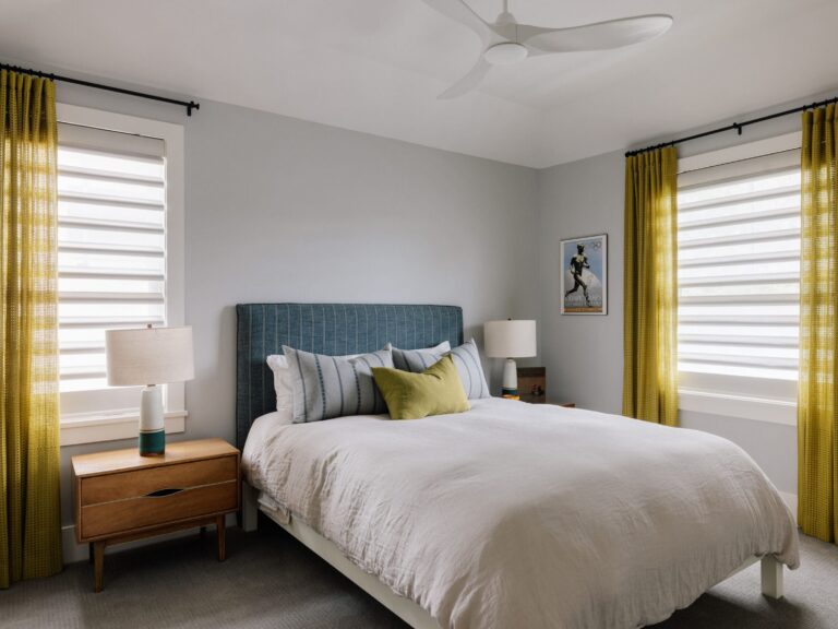 A modern bedroom with a blue upholstered bed, white bedding, two wooden nightstands with lamps, yellow curtains, and two large windows.