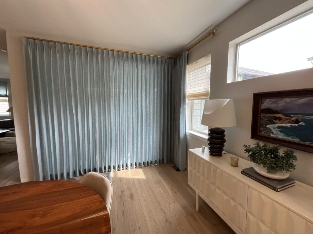 A dining area with a round wooden table, blue floor-length curtains, a white sideboard, lamp, plant, and a seaside painting under a window with blinds.