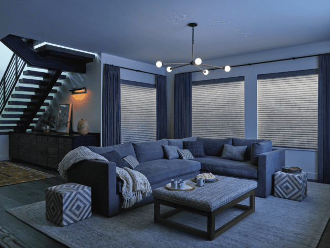 Living room with a large gray sectional sofa, an upholstered ottoman, and windows covered by pleated shades.