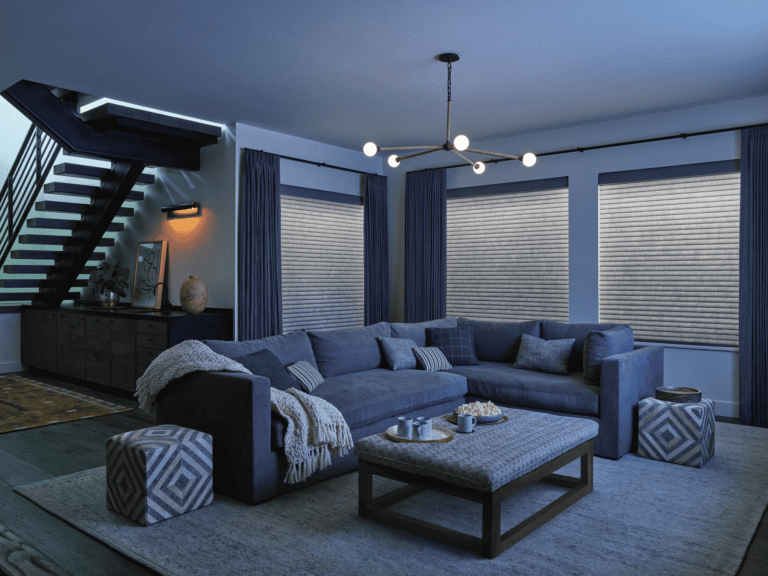 Living room with a large gray sectional sofa, an upholstered ottoman, and windows covered by pleated shades.